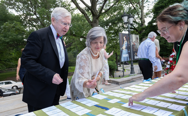 An older white couple in fancy dress attire, standing in front of a table with a white woman in all black assisting them.