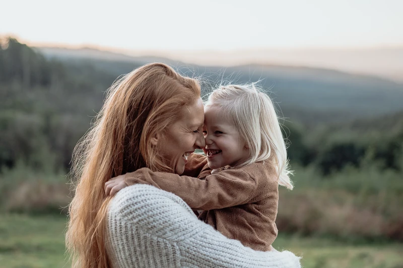 A mother hugging her 3 year old daughter, resting their foreheads on each other, cheeky smiles on their faces.