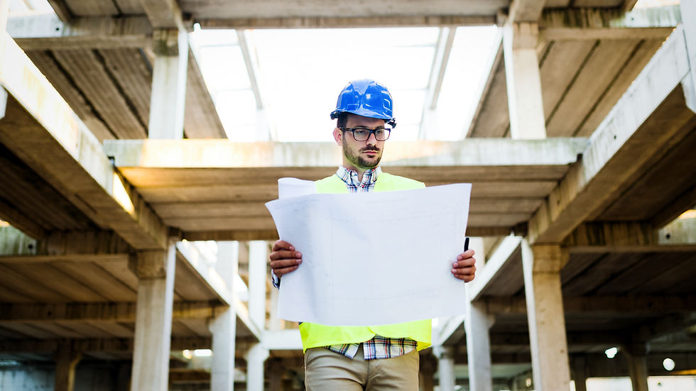 Construction worker in a blue helmet and yellow vest studies blueprints inside an unfinished building. Bright daylight enters through open beams.