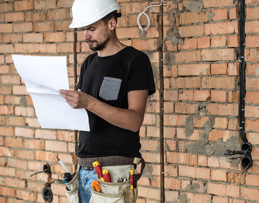 Construction worker in a white hard hat reads a paper. He wears a tool belt and black shirt, standing against a brick wall.