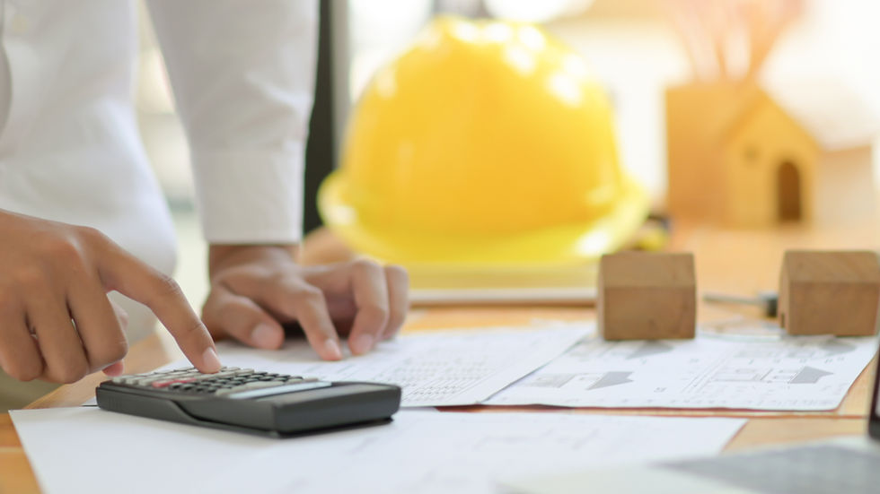 Hands using a calculator on architectural plans. A yellow hard hat and small wooden models in the background. Bright, professional setting.