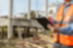 Construction worker in orange vest holds tablet, inspecting building site with concrete structure and scattered wood. Bright, sunny day.