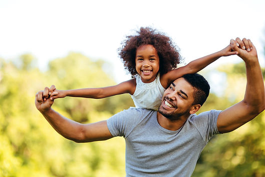 A man lifts a young girl on his back while both smile with thier arms out wide.