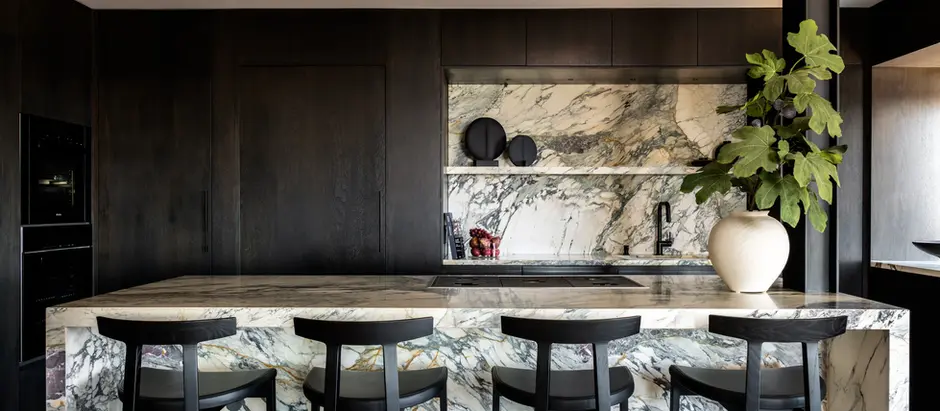 Modern kitchen with marble counters, sleek black chairs, and dark cabinets. A large plant in a white vase adds a touch of greenery.
