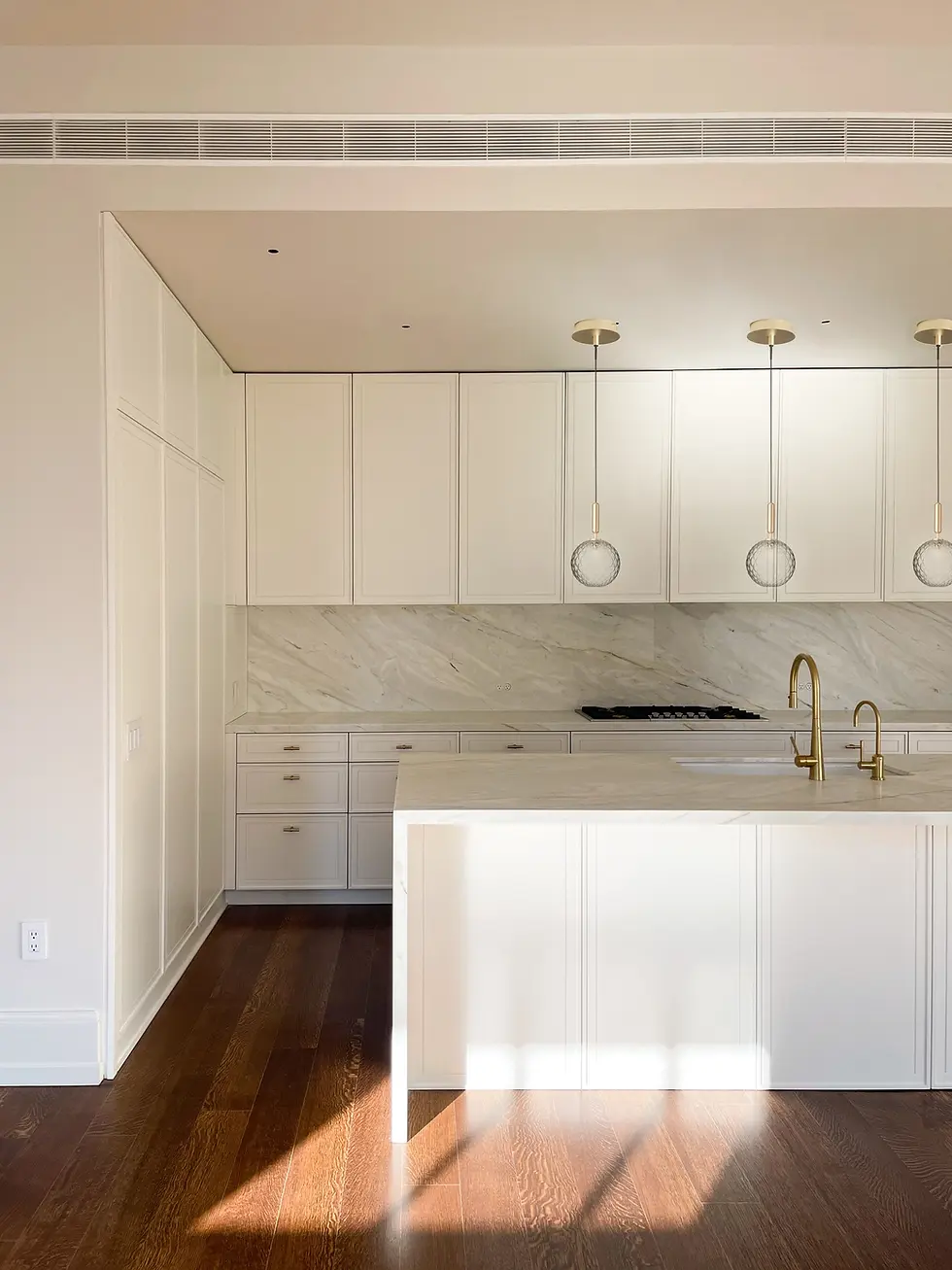 Bright kitchen with white cabinets, marble backsplash, and island. Three pendant lights hang above, golden faucets, and wooden flooring.