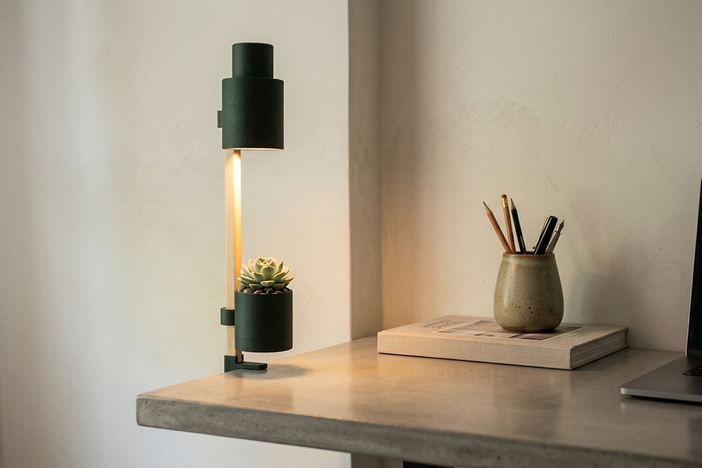 A stylish Folt Modular lighting and planter unit in a cozy, minimalist interior. The design features a white cylindrical lamp at the top and vertically stacked pots housing various succulents and cacti at the base, positioned next to a wooden lounge chair with a soft throw blanket.