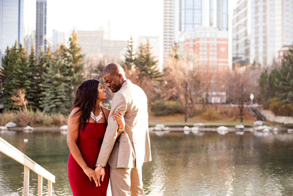 Beautiful couple, him in a suit and she is a red dress in front of a small lake with a city scape in the background.