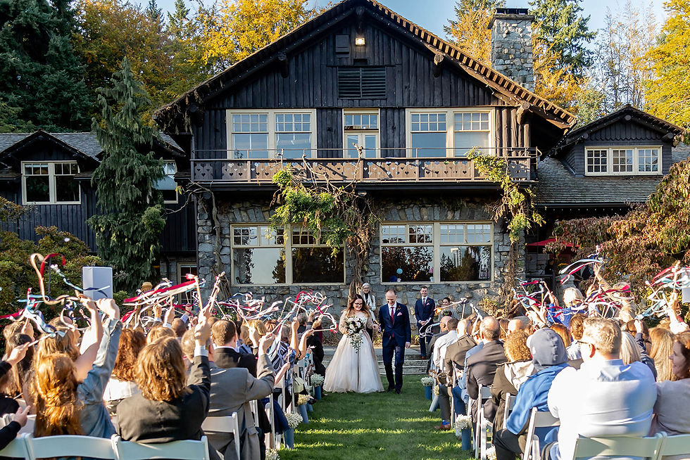 Outdoor wedding ceremony at the Stanley Park pavillion in Vancouver. The bride and groom are walking up the aisle and the guests are all waving ribbons.