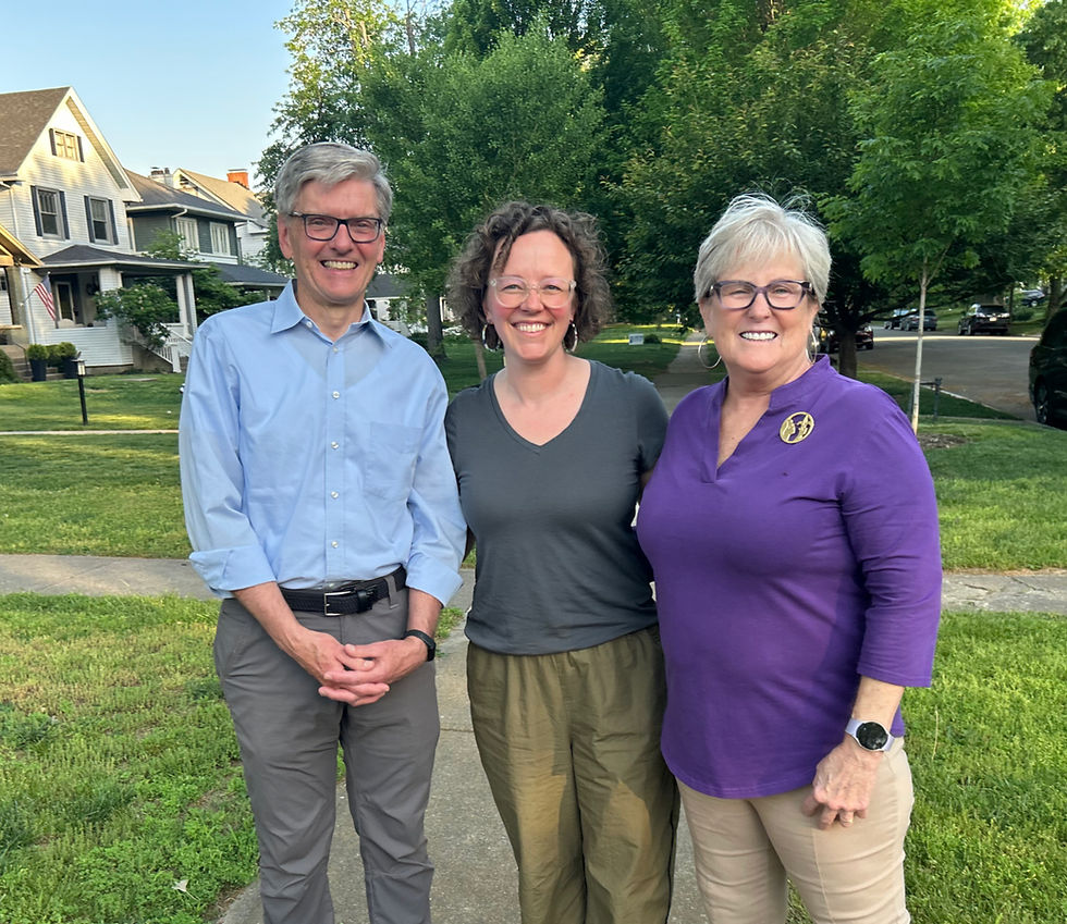 Three people smiling outdoors on a sunny day, standing on a sidewalk in front of houses. One, Tina Ward-Pugh, wears glasses and a purple shirt with a pin. Alison Brotzge-Elder is wearing clear glasses, multicolored hoop earrings and has curly hair. She's wearing a gray shirt and green pants. Bill Hollander is wearing glasses, a blue button down shirt, and gray pants.