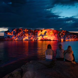 Three women sitting on a rock looking across the sea at a huge granite rock lit up with artwork