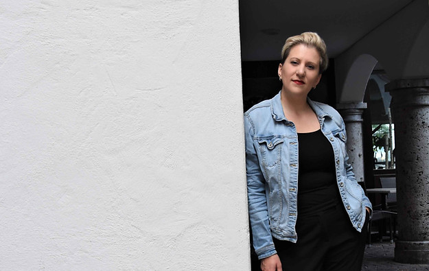 Christianne Bélanger lehnt in Jeansjacke an einer weißen Wand, Arkaden im Hintergrund.