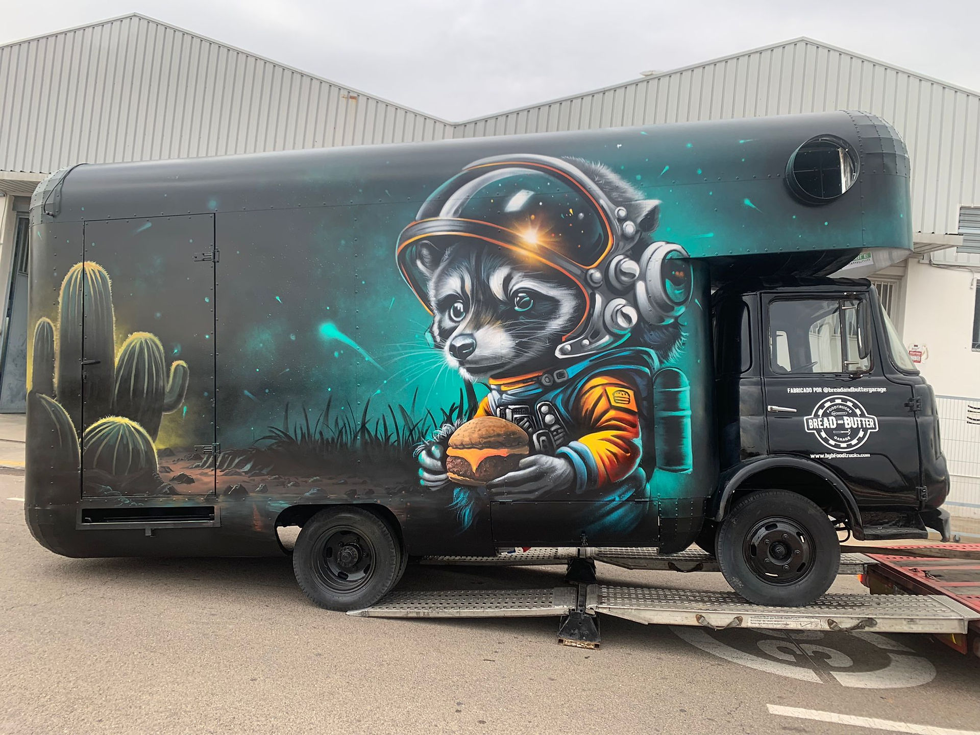 Food truck negro con grafiti de mapache astronauta comiendo hamburguesa.
