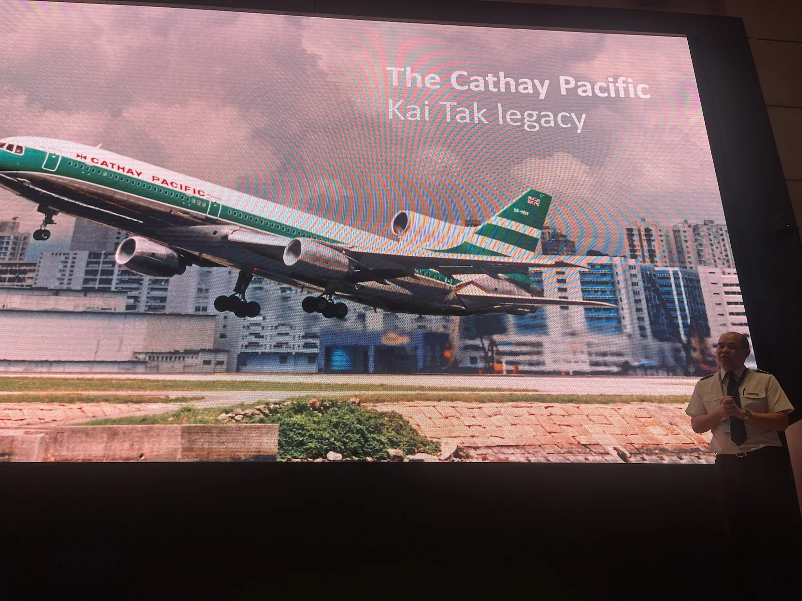 A captain speaks beside a presentation slide showing a Cathay Pacific Boeing 747 and the words The Cathay Pacific Kai Tak legacy.