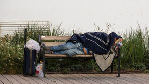 A homeless person sleeping on a public bench under a blanket with a suitcase and plastic bags next to the bench