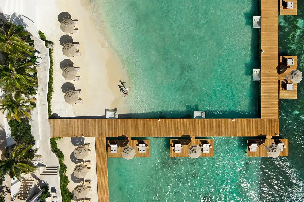 Aerial view of a beach with turquoise water, wooden piers, and patio umbrellas. Two people walk along the shoreline. Calm and serene at Impression by Secrets Isla Mujures