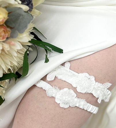 Bride wearing crepe white satin dress with high slit and holding ivory bouquet. Pale leg is showing wearing two lace flower custom bridal garters made from alteration scraps