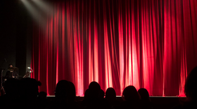 Red stage curtains under a spotlight with audience silhouettes in front