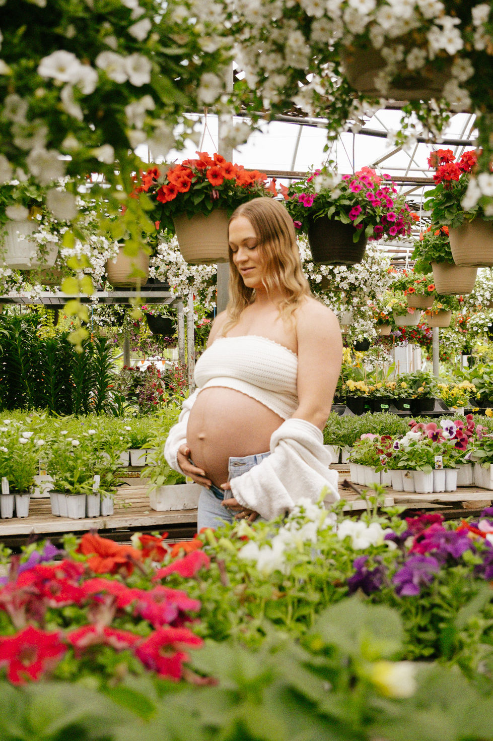 Motherhood maternity photo with expectant mother surrounded by florals and greenery, soft natural light creating a dreamy, serene atmosphere.