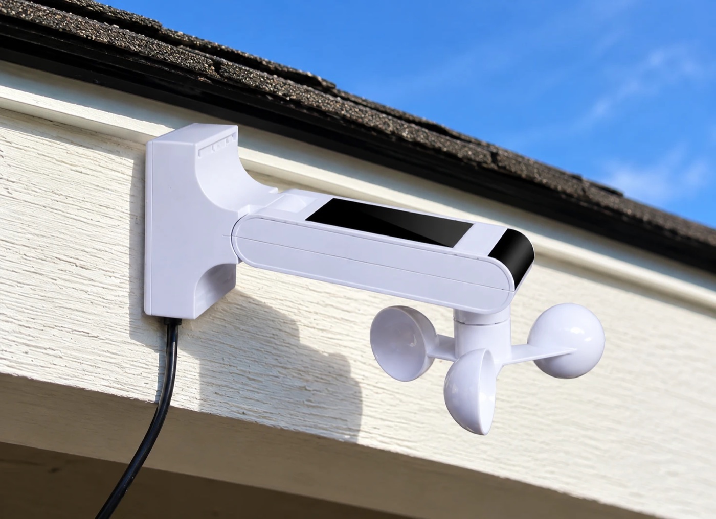 The white wind and rain sensor mounted under the eave of a house, with its three spinning cups and solar panel