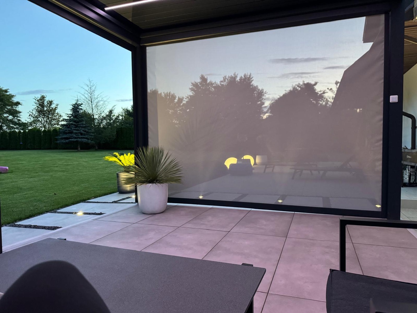 A retractable zip screen lowered on one side of a black pergola at dusk