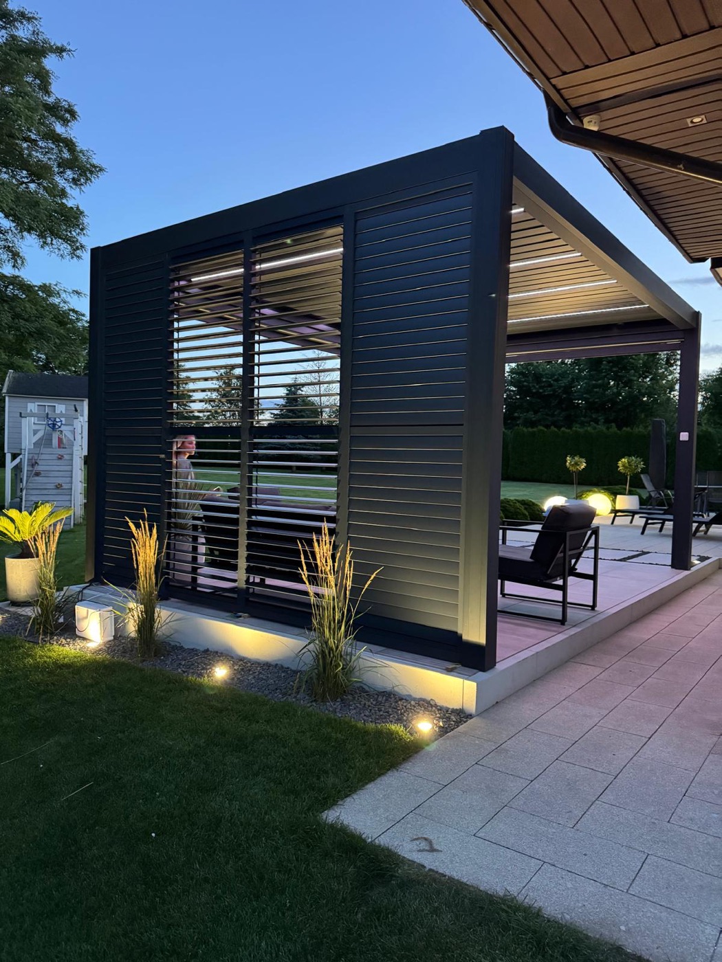 Aluminum louver shutter panels closing one side of a black pergola, lit from below