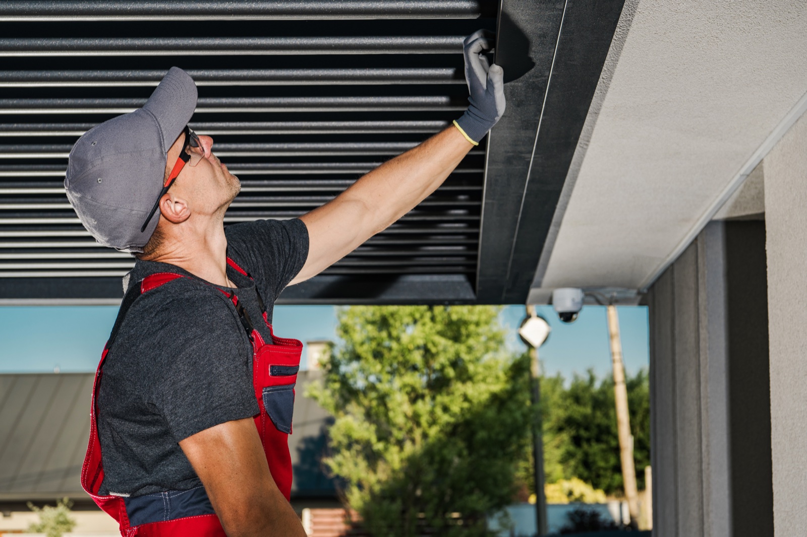 An installer in red work overalls fitting a black louvered pergola roof against a home