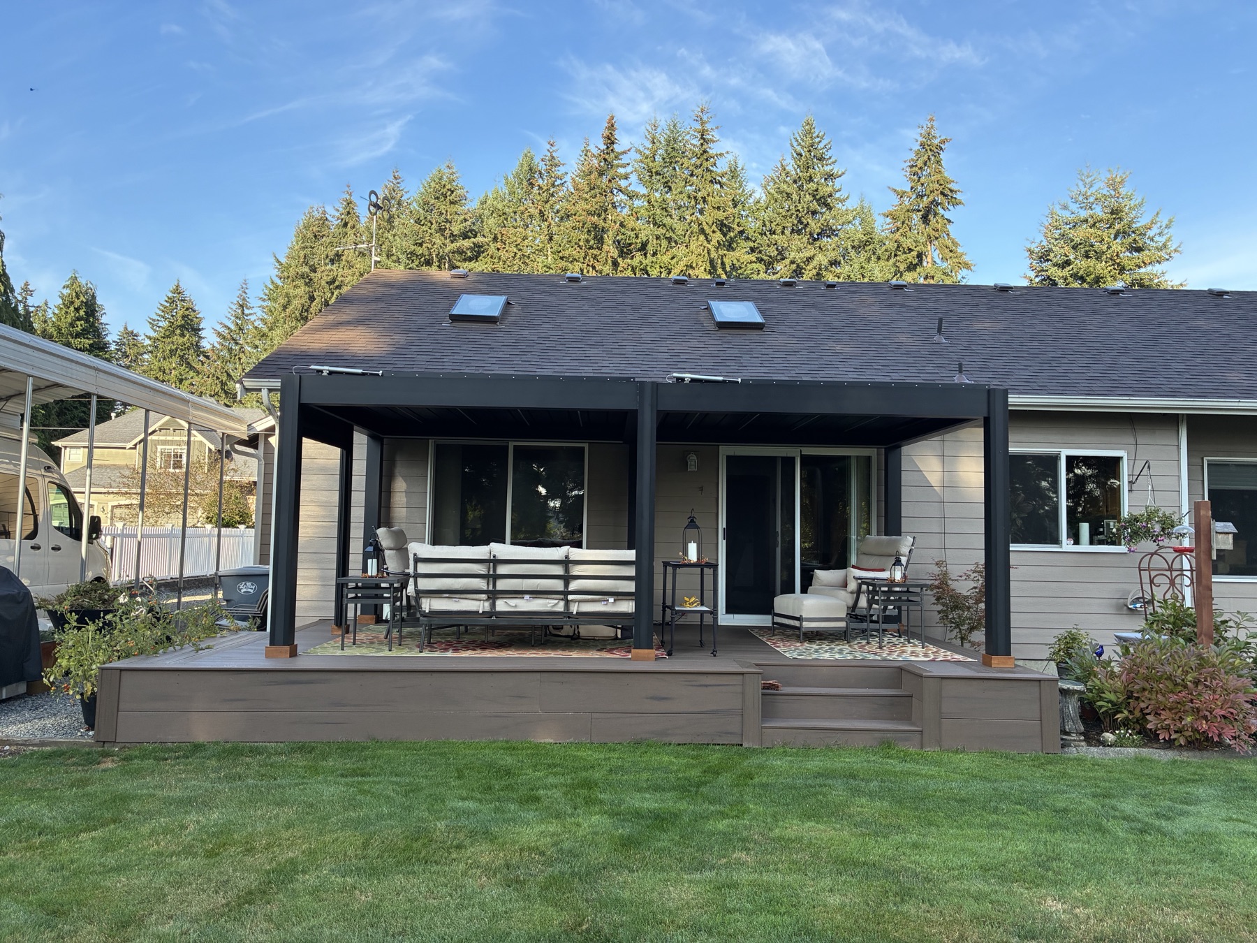 A black louvered pergola over a raised composite deck behind a grey single-story home in Puyallup, with evergreens behind