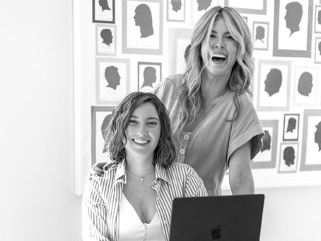 Gesche and Christina smiling at a desk with an open laptop, posing before a wall of colorful silhouette portraits.