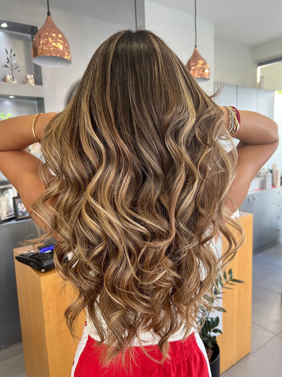 Woman with long, wavy, highlighted hair stands in a salon, hands in hair. Copper pendant lights and modern decor in the background.