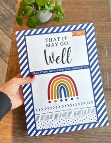 Rainbow Homeschool book from That It May Go Well Free Christian Homeschool Curriculum by Lamp and Light Living Sitting on a table with a plant in the background.