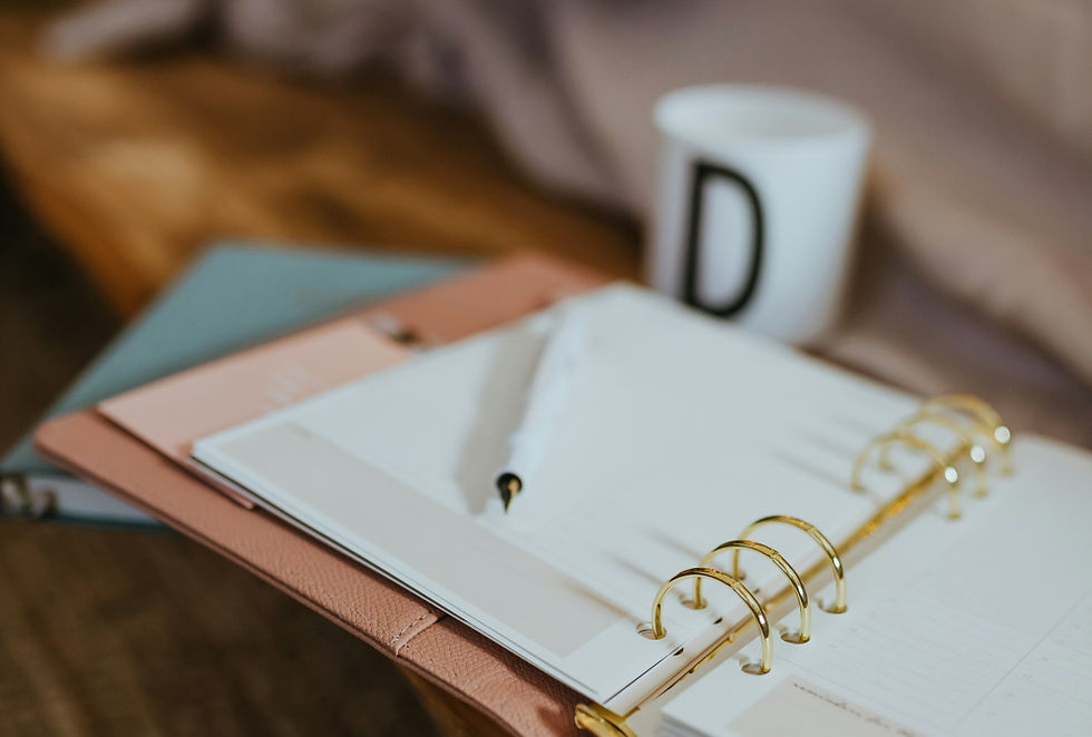 A close up photo of an open spiral bound notebook with a white pen sitting on top. There is a white mug with a black letter D on it, slightly blurred in the background.