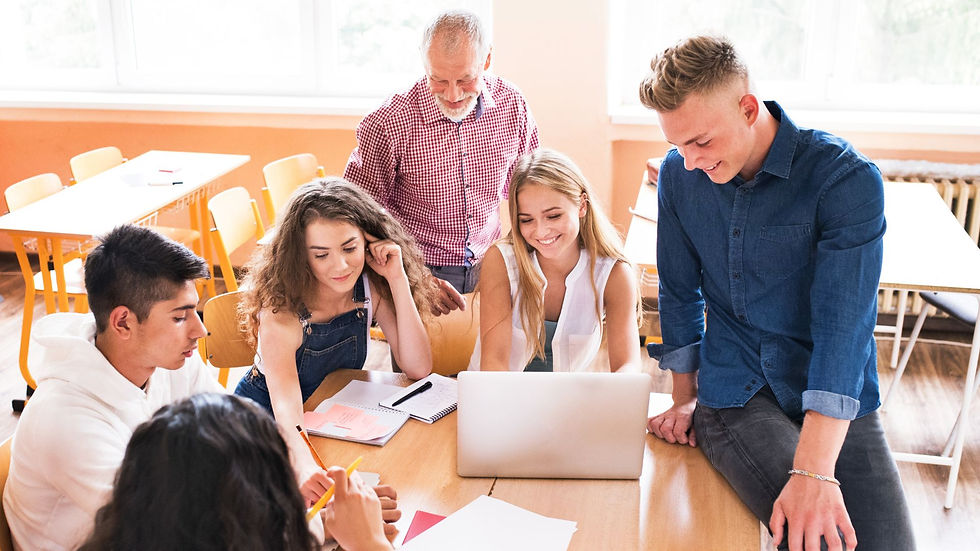 A small group of high school students work together in a classroom with a male teacher while using pen, paper, and a lap top.