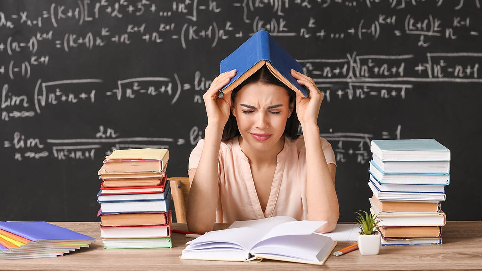 A lady teacher sits at her desk in front of a blackboard full of math formulas. She has books around her, and is holding a blue book on her head in despair.
