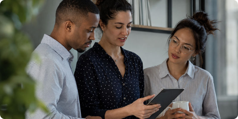 Three coworkers in an office review a tablet together, one holding coffee, with a calm, focused mood.