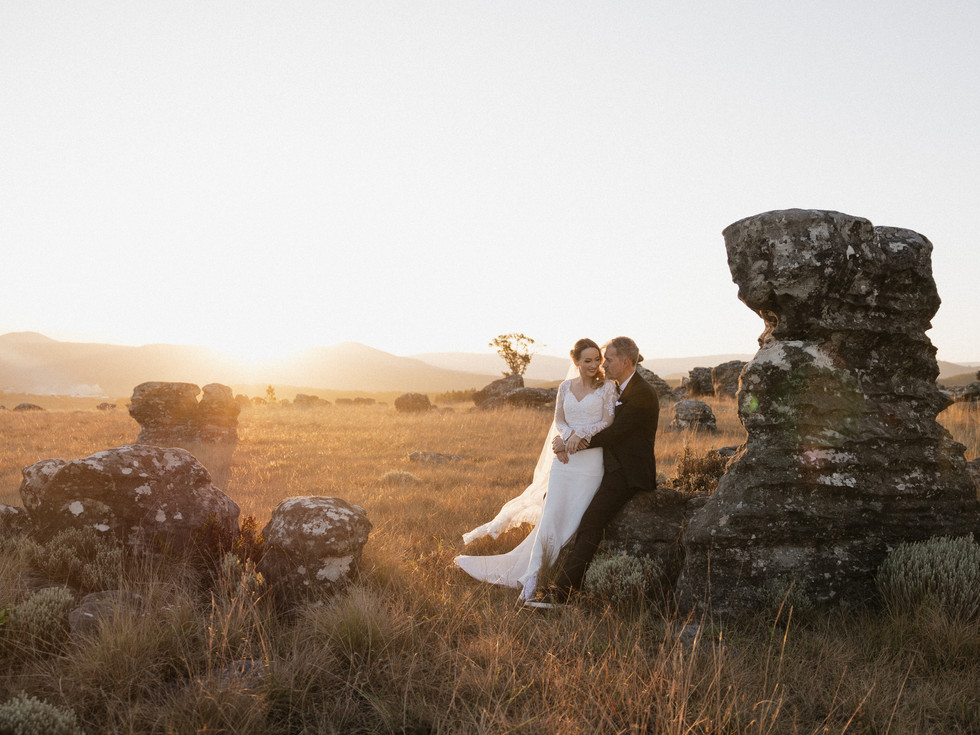 Bride and groom embrace while sitting on rocks in golden sunset landscape during South Africa helicopter elopement