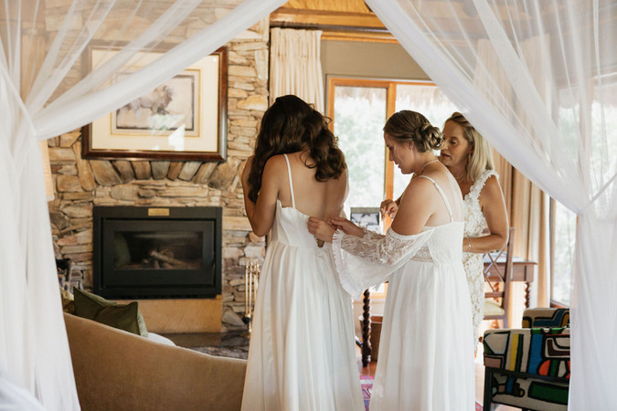 Bride in white gown, assisted by two women adjusting dress in cozy room with stone fireplace, curtains, and soft natural light.