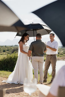 Bride and groom under a black umbrella during an outdoor Safari Wedding ceremony in Hoedspruit South Africa. Celebrant reads in bright, natural setting with mountains in background.