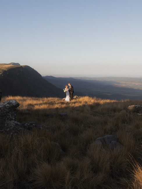 Bride and groom standing together on grassy cliff edge overlooking Panorama Route mountains during helicopter elopement in South Africa
