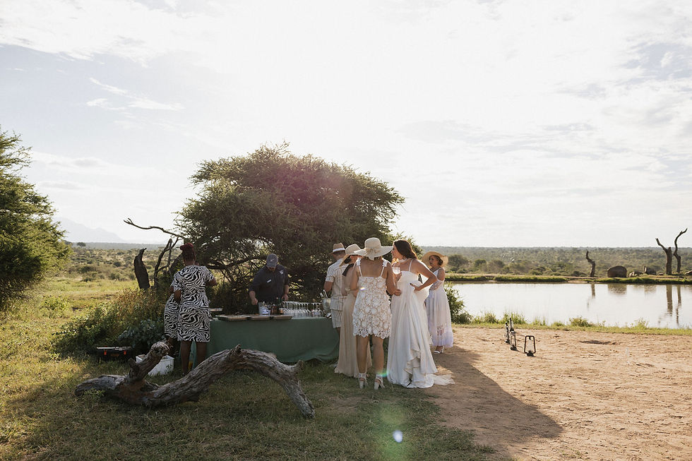 People gather around a table outdoors near a dam, dressed in light, summery attire. Sun shines through trees, creating a serene atmosphere.