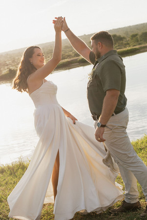 A couple joyfully dances by a sunlit dam. The woman wears a flowing white dress; the man is in a green shirt. Background is serene nature.
