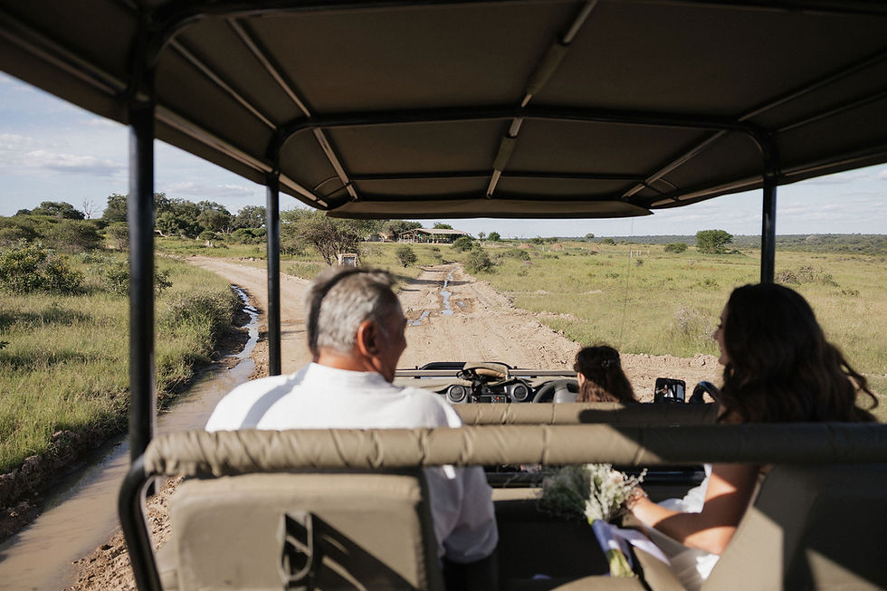 Safari vehicle with bride and father drives on a dirt road in a grassy savanna. Clear sky, relaxed mood, bouquet visible in the foreground.