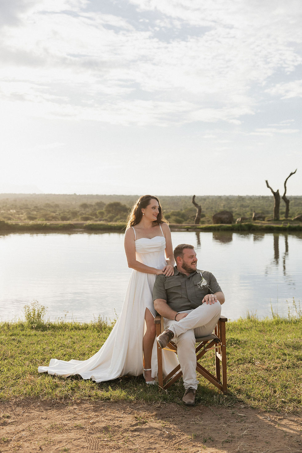 A woman in a white dress stands by a seated man on a wooden chair. They're by a dam with a scenic view, under a partly cloudy sky.