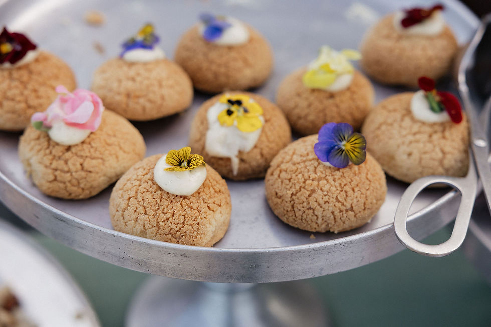 Cream puffs topped with colorful edible flowers on a silver tray. The background is blurred, and a serving tong is visible.