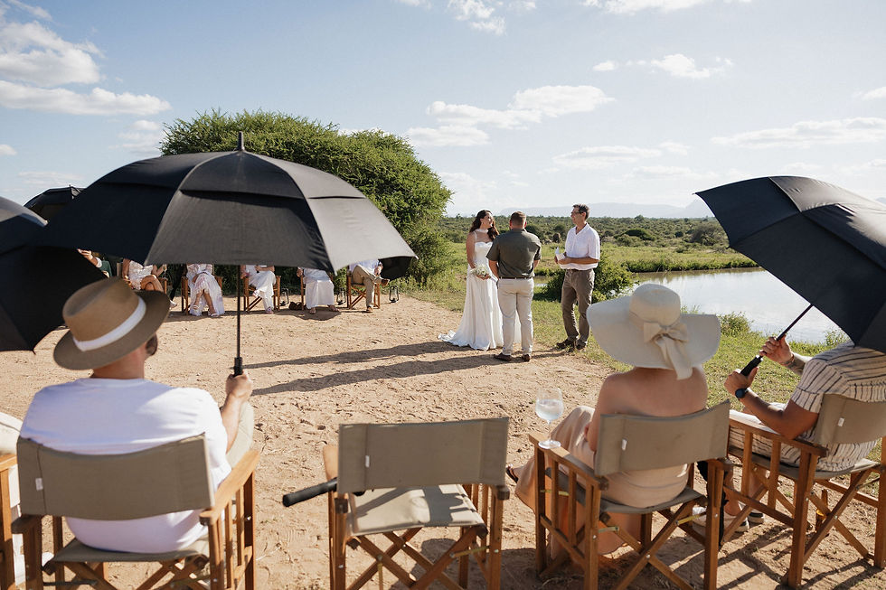 Outdoor wedding with a couple exchanging vows near a dam. Guests sit under umbrellas in chairs. Clear sky and lush greenery surround them.