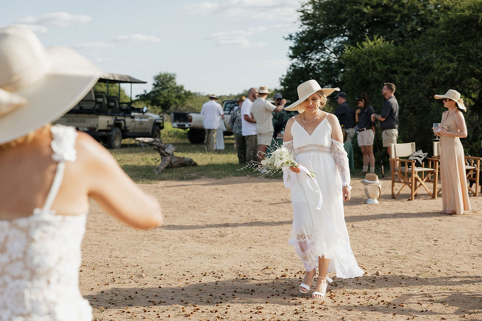 Woman in white dress and hat walks on sandy path holding flowers. Safari vehicles and people in background. Sunny outdoor setting.