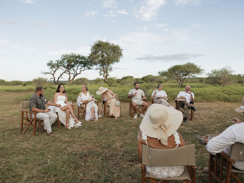 A group of people sits in a circle of chairs outdoors on a grassy safari clearing under a blue sky, with trees in the background. They're dressed in light, summery attire.