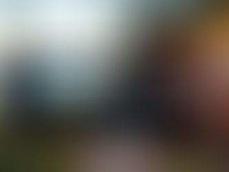 A hand holds a smartphone horizontally, recording a video of a young man sitting by a scenic lake at sunset. The phone's screen displays video recording controls and settings, with blurred string lights in the background.