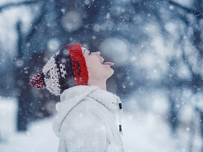 A person in a white coat and colorful hat catches snowflakes on their tongue in a snowy woodland setting, conveying joy and wonder.