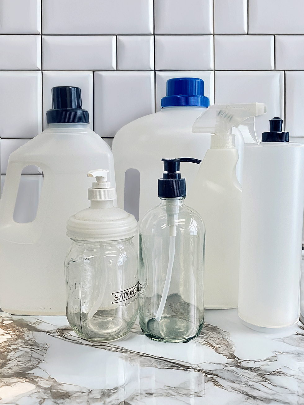 Assorted clear and white containers with pumps and caps on a marble counter. White tiled wall background. Text "SAPONETTI" visible.