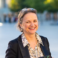 Trois femmes sourient sur la place, tenant des documents, les arbres en arrière-plan.